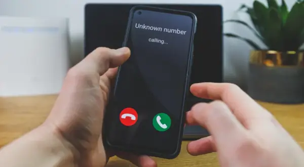 Dłoń trzyma smartfon, na ekranie którego widnieje komunikat "Unknown number calling...". Numer prywatny dzwoni, a użytkownik zastanawia się, czy odebrać.