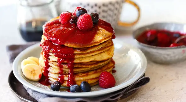 Ein Stapel goldbrauner Dinkel Pancakes, übergossen mit roter Beerensauce und garniert mit frischen Beeren und Bananenscheiben.
