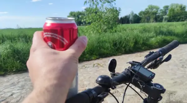 Rowerzysta trzyma puszkę napoju, planując jazdę rowerem po alkoholu. W tle zielone pola i błękitne niebo.