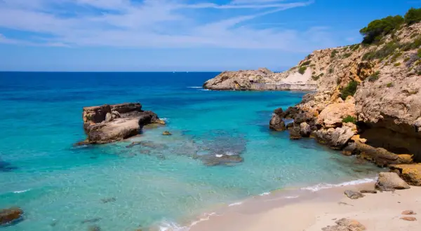 Aguas cristalinas y rocas escarpadas en una cala idílica. Unos pequeños **planes** se ven en el horizonte, invitando a soñar con esta **playa** paradisíaca.