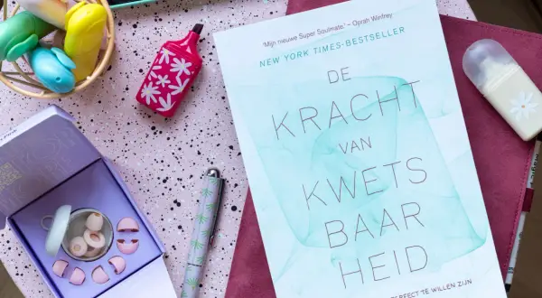 Boek 'De Kracht van Kwetsbaarheid' en oordopjes, ideaal voor wie last heeft van omgevingsgeluid. Creatieve spullen op een roze ondergrond.