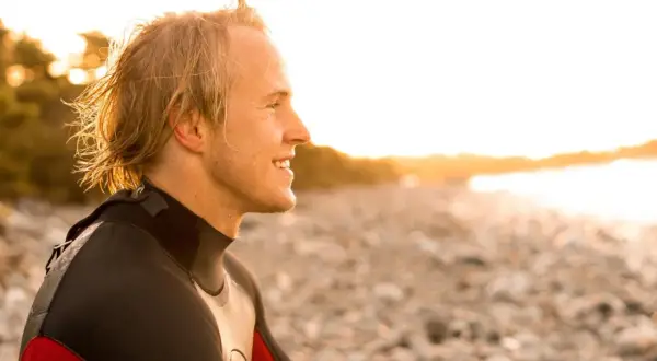 Uśmiechnięty surfer w piance, na planie średnim, patrzy w stronę zachodzącego słońca na kamienistej plaży.