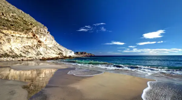 Playa de arena dorada y acantilados rocosos en la cala del plomo, con el mar azul intenso y olas espumosas.