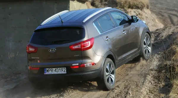 Brązowy Kia Sportage 2014 zjeżdża po piaszczystej ścieżce, unosząc kurz.