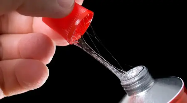 Main d'une personne tenant le bouchon rouge d'un tube de colle super glue, avec des fils de colle s'étirant. Attention, colle super glue sur les doigts dangereux.