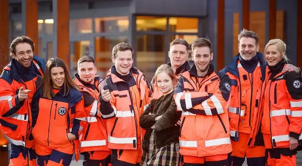 Uśmiechnięta obsada serialu "Na sygnale" w pomarańczowych kurtkach ratowniczych pozuje przed budynkiem.