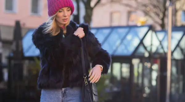 Krótkie futerko stylizacje: blondynka w różowej czapce i jeansach, z czarną torebką.