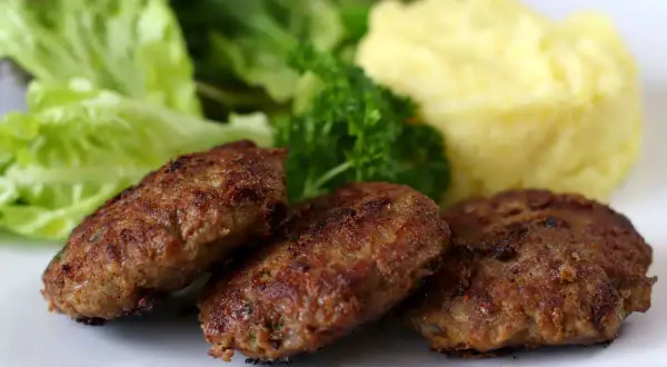 Drei goldbraune bayerische Fleischpflanzerl liegen auf einem Teller neben cremigem Kartoffelpüree und frischem Salat.