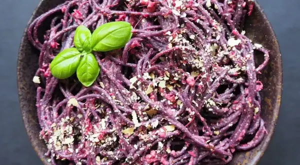 Fioletowy makaron z pesto z buraków, posypany orzechami i udekorowany listkami bazylii.