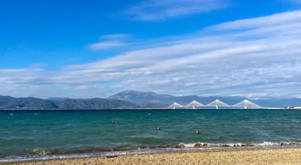 Spokojne morze i plaża w Grecji w styczniu. W oddali widać most i góry.