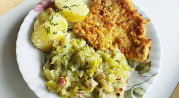 Kotlet schabowy z ziemniakami i zasmażaną kapustą z boczkiem na talerzu.