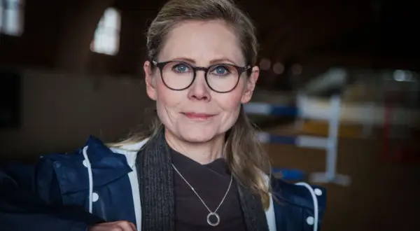 Anna Schreil med glasögon och en blå jacka, står i en ridhall.