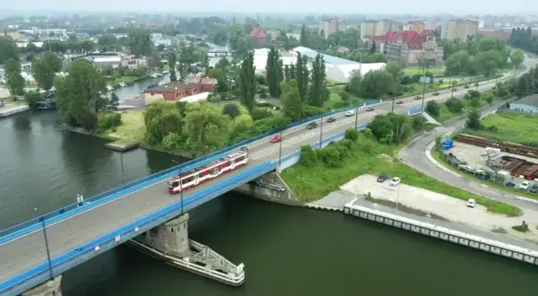 Tram z czerwonymi pasami jedzie przez most nad rzeką, w tle widać miasto i tereny przemysłowe, jak na plaży Stogi.