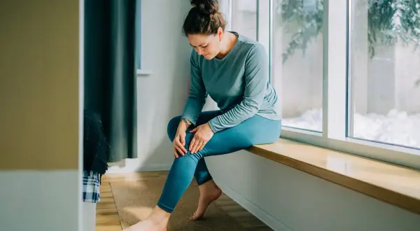 Femme assise près d'une fenêtre, tenant son genou, possiblement à cause d'une **durée courbature** après un effort.