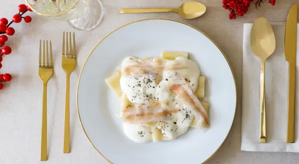 Piatto di paccheri conditi con una cremosa spuma di pecorino e guarniti con pepe nero. Accanto, posate dorate e un rametto di bacche rosse.