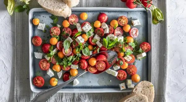 Frischer Tomate Mozzarella Salat mit bunten Kirschtomaten, Feta und Minze, serviert auf einem grauen Teller mit Brot.