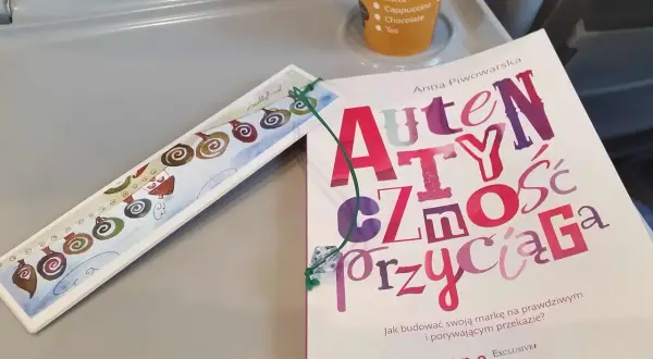 Książka "Autentyczność przyciąga" Anny Piwowarskiej na stoliku, obok ozdobna zakładka i kubek z napojami.