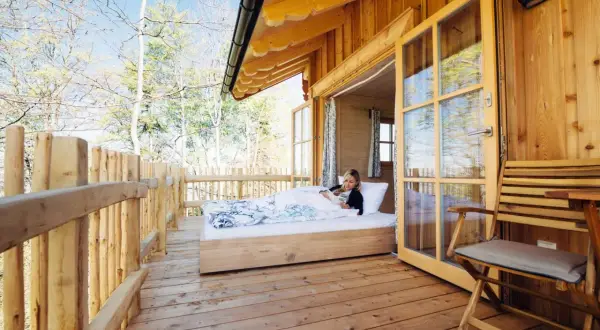 Frau liest im Bett in einer Holzhütte mit offener Tür und Blick auf Bäume.