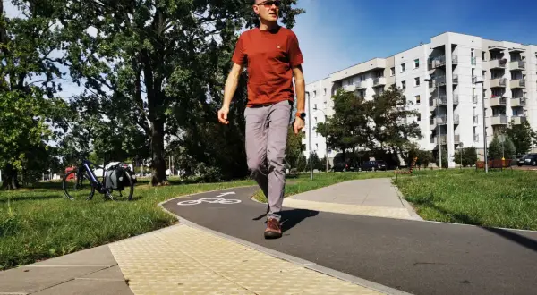 Mężczyzna idzie ścieżką rowerową, na której widnieje symbol roweru. Obok ścieżki znajduje się chodnik z wypukłą nawierzchnią, sugerujący przejście dla pieszych.