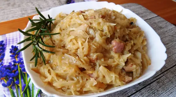 Pyszna kapusta zasmażana z boczkiem, podana na białym talerzu, ozdobiona gałązką rozmarynu.