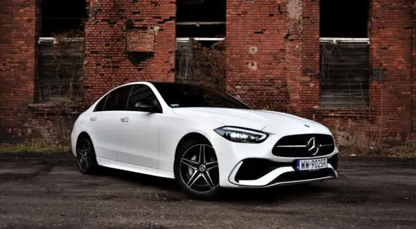 Biały Mercedes 220 z czarnym dachem zaparkowany przed zniszczoną ceglaną ścianą.