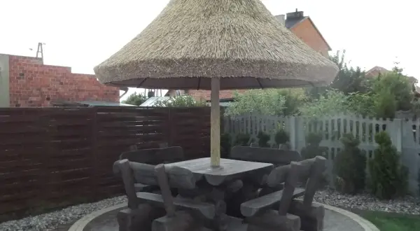 Drewniany stół z ławami pod dużym, słomianym parasolem ogrodowym.