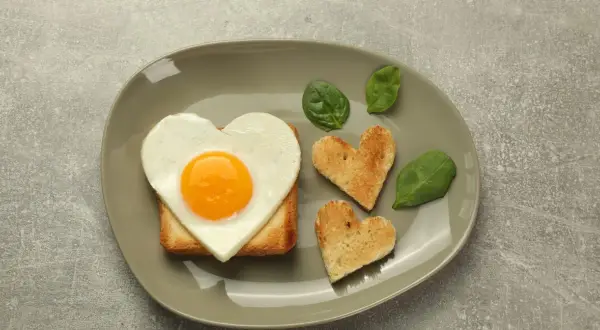 Jajko sadzone w kształcie serca na grzance, obok tostów w kształcie serc i liści szpinaku.