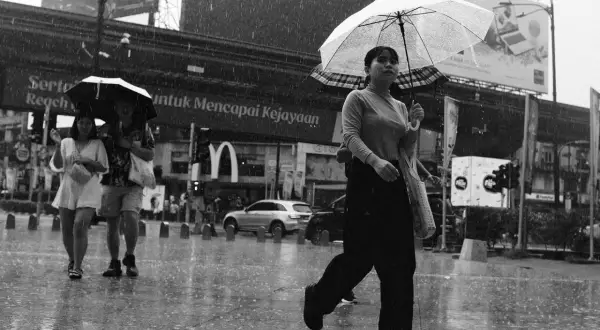 A woman with a clear umbrella walks in the rain, a tri x sign visible in the background.