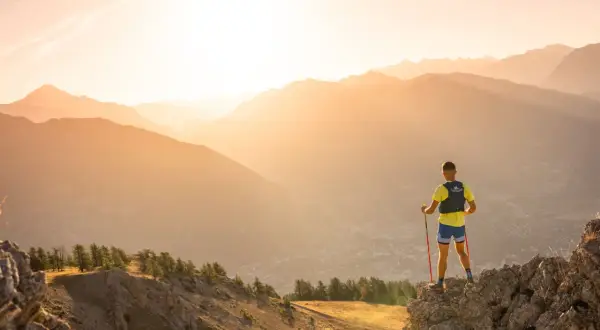 Un coureur de trail, bâtons en main, admire le paysage montagneux au lever du soleil.