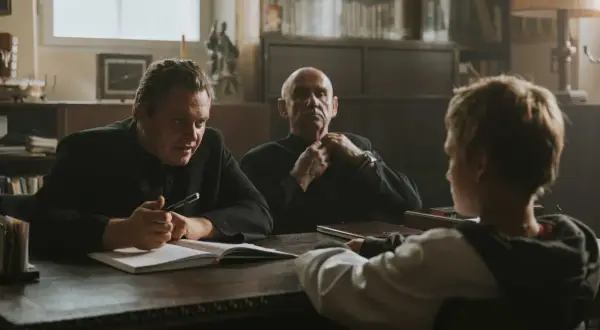 Trzech mężczyzn przy stole, jeden z nich pisze. Scena z filmów Piotra Domalewskiego.