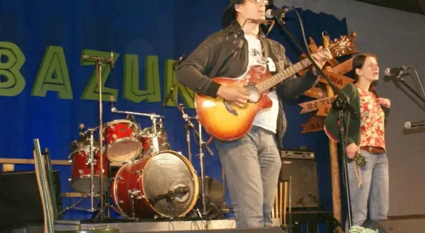 Rafał Chojnacki gra na gitarze i harmonijce ustnej, obok niego śpiewa kobieta. W tle zespół muzyczny i niebieska kurtyna z napisem "BAZUNA".