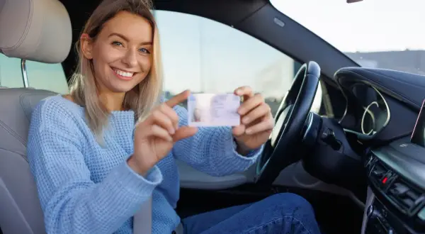 Uśmiechnięta kobieta w niebieskim swetrze pokazuje swoje nowe zdjęcie do prawa jazdy, siedząc za kierownicą samochodu.