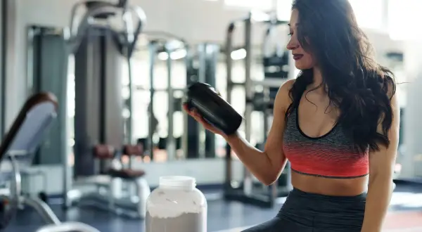Frau im Fitnessstudio hält einen Shaker, daneben ein großer Behälter mit Pulver. Perfekt für den **proteinshake vor oder nach dem training**.
