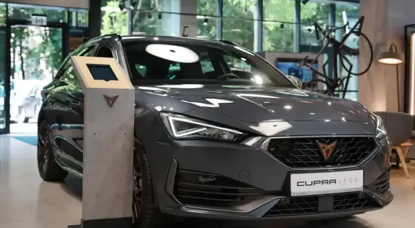 Nowoczesne konie mechaniczne, szary Cupra Leon, prezentują się w salonie. Obok stoi multimedialny kiosk.