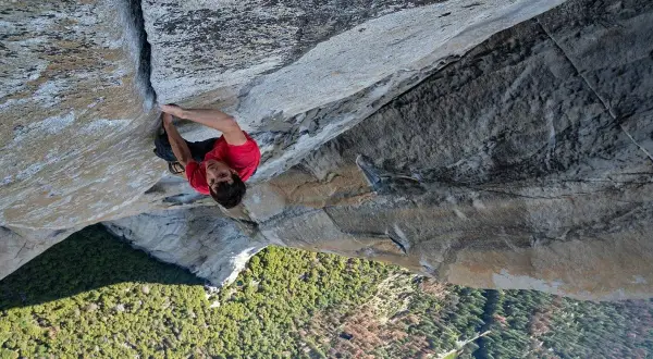 Alex Honnold klettert free solo die steile Felswand hinauf. Unter ihm erstreckt sich ein grünes Tal mit Bäumen.