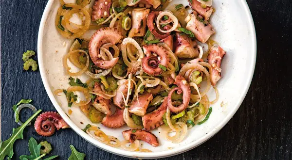 Un delicioso salpicón de pulpo con cebolla y perejil, servido en un plato blanco sobre una pizarra oscura.