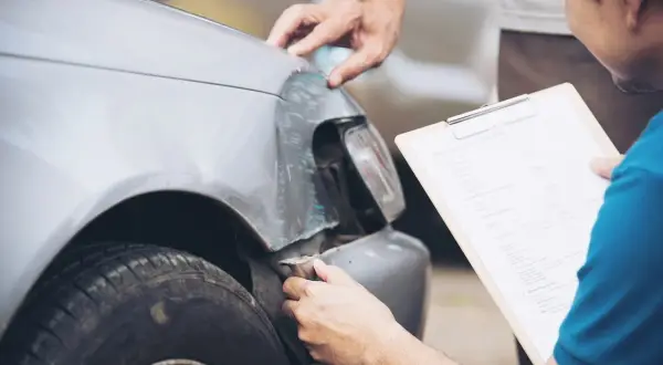 Deux hommes examinent les dégâts sur une voiture, peut-être suite à un délit de fuite. L'un tient un formulaire de déclaration de sinistre.