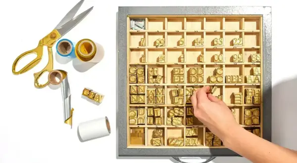 A hand selects a letter stamp from a tray of vintage letterpress type, part of a collection for creating a monogram.