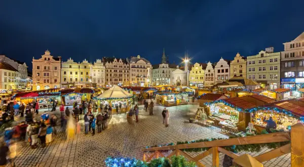 Beleuchtete Buden und historische Gebäude auf einem tschechischen Weihnachtsmarkt. Menschen schlendern über den Platz, der mit Lichterketten geschmückt ist.