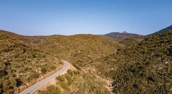 Vue aérienne d'une route sinueuse traversant des collines verdoyantes, menant vers le col du Testanier. Deux cyclistes s'engagent sur la pente.