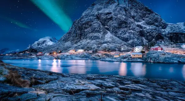 Grüne Bänder der Aurora Borealis tanzen über einem verschneiten Berg, der eine kleine Küstensiedlung mit roten und gelben Häusern beleuchtet.