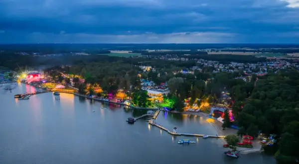 Nocny widok na jezioro i rozświetlone tereny imprezowe. Widać scenę, tłumy ludzi i kolorowe światła, które tworzą atmosferę sława festiwal.