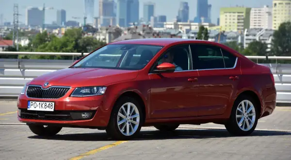 Czerwona Skoda Octavia 3 z widocznymi żarówkami w przednich reflektorach, zaparkowana na tle miasta.