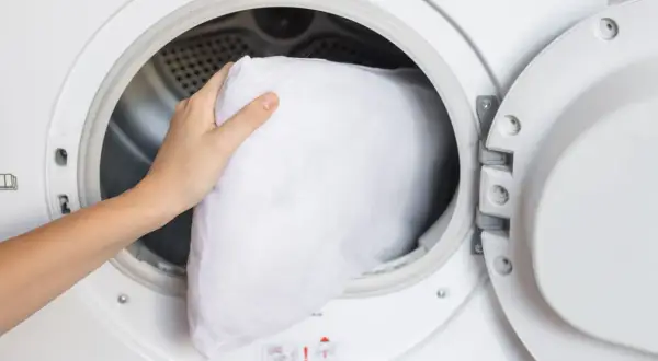 A hand places a white pillow into a washing machine, demonstrating how to wash pillows in a washing machine for a fresh, clean sleep.
