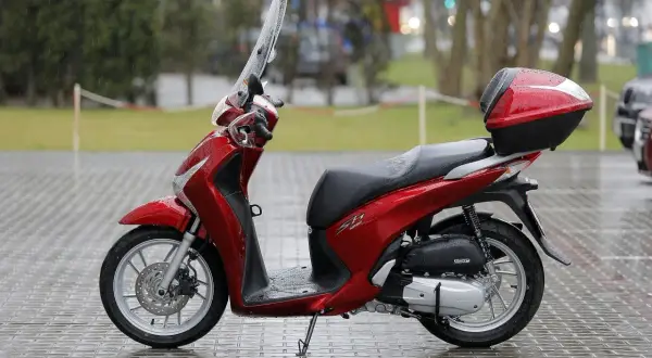 Czerwony skuter Honda SH z kufrem, gotowy do jazdy. W Polsce od ilu lat można jeździć skuterem? Od 14 lat na 50cc, od 18 lat na większe pojemności.