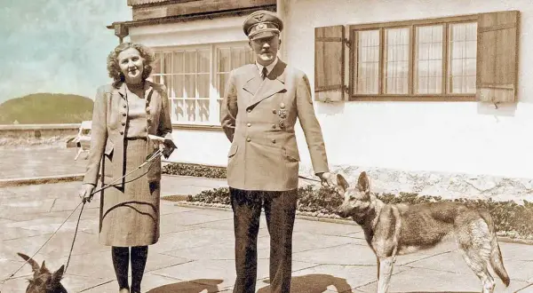 Eva Braun, hitlers fru, står med en hund. Bredvid henne står en man i uniform med en annan hund.