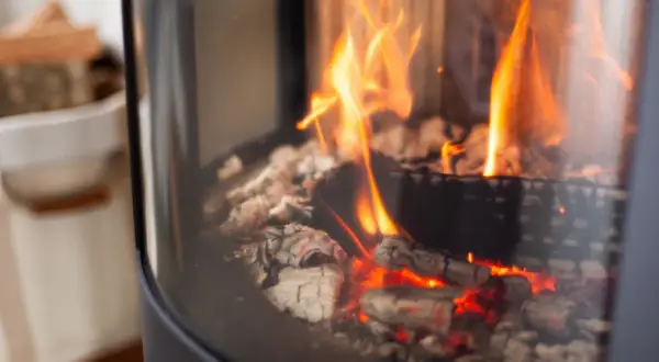 Glühende Kohlen und Flammen in einem modernen Kaminofen. Links daneben ein Korb mit Holzscheiten.