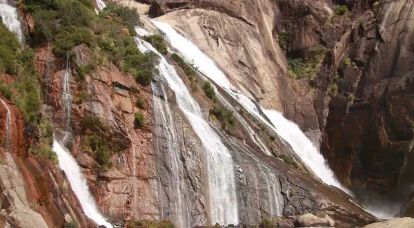 Cascada de agua blanca cayendo por rocas rojizas y grises, rodeada de vegetación. Un espectáculo natural de las fervenzas de Galicia.