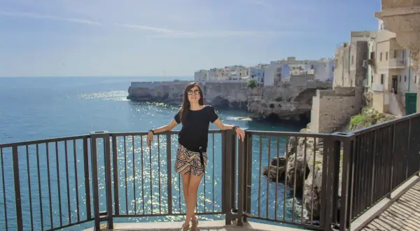 Donna sorridente con occhiali da sole si appoggia a una ringhiera con vista sul mare cristallino e le case bianche di Polignano a mare.