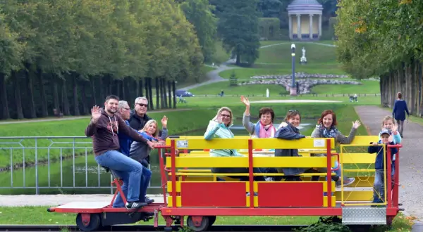 Fröhliche Menschen genießen eine Draisinenfahrt NRW durch eine Parklandschaft mit einem historischen Gebäude im Hintergrund.